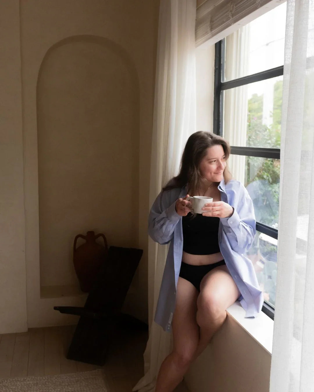 Woman in black period underwear and shirt enjoying tea by a window, relaxed indoor scene