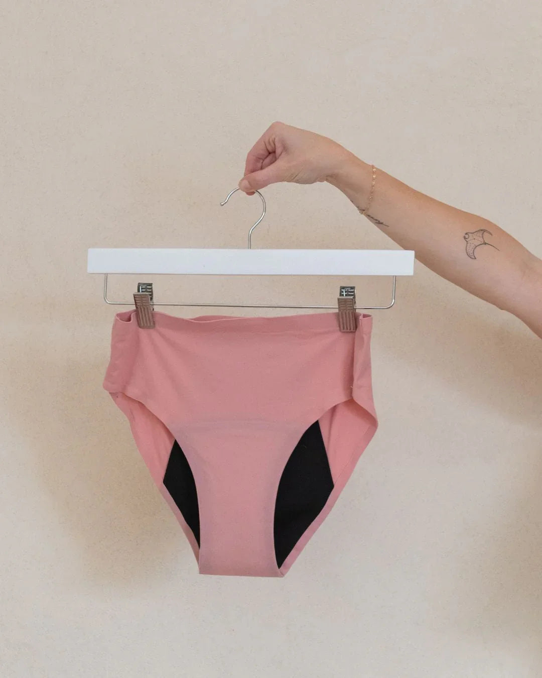 Hand holding a hanger with pink leak-proof period underwear against a beige background.