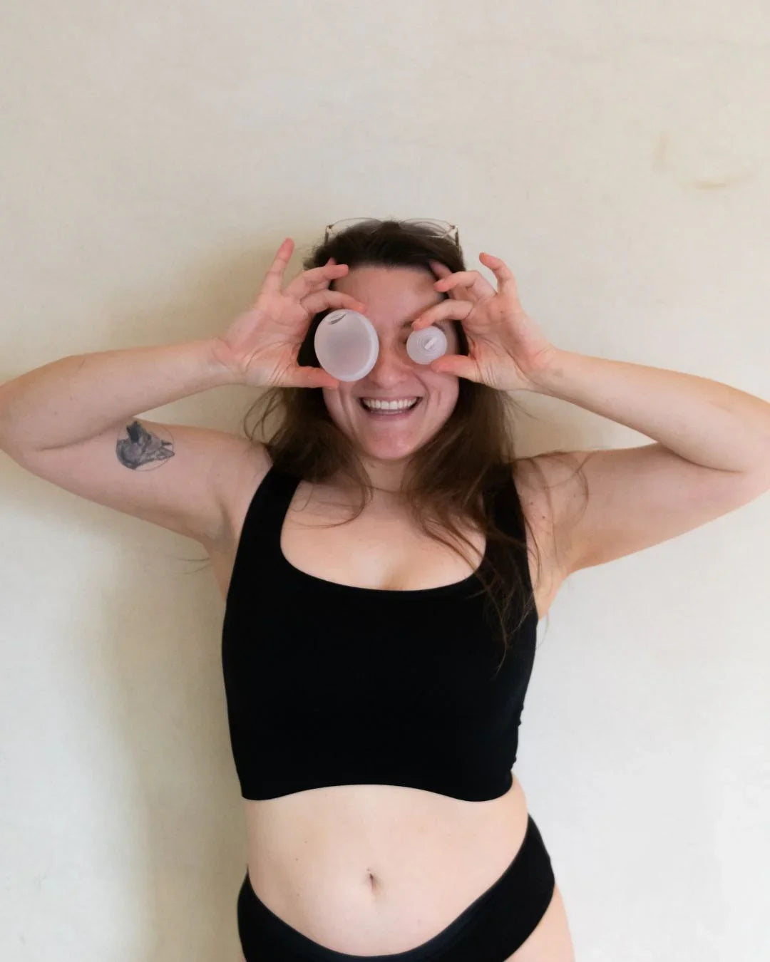 Smiling woman in black period underwear holding menstrual cups over her eyes