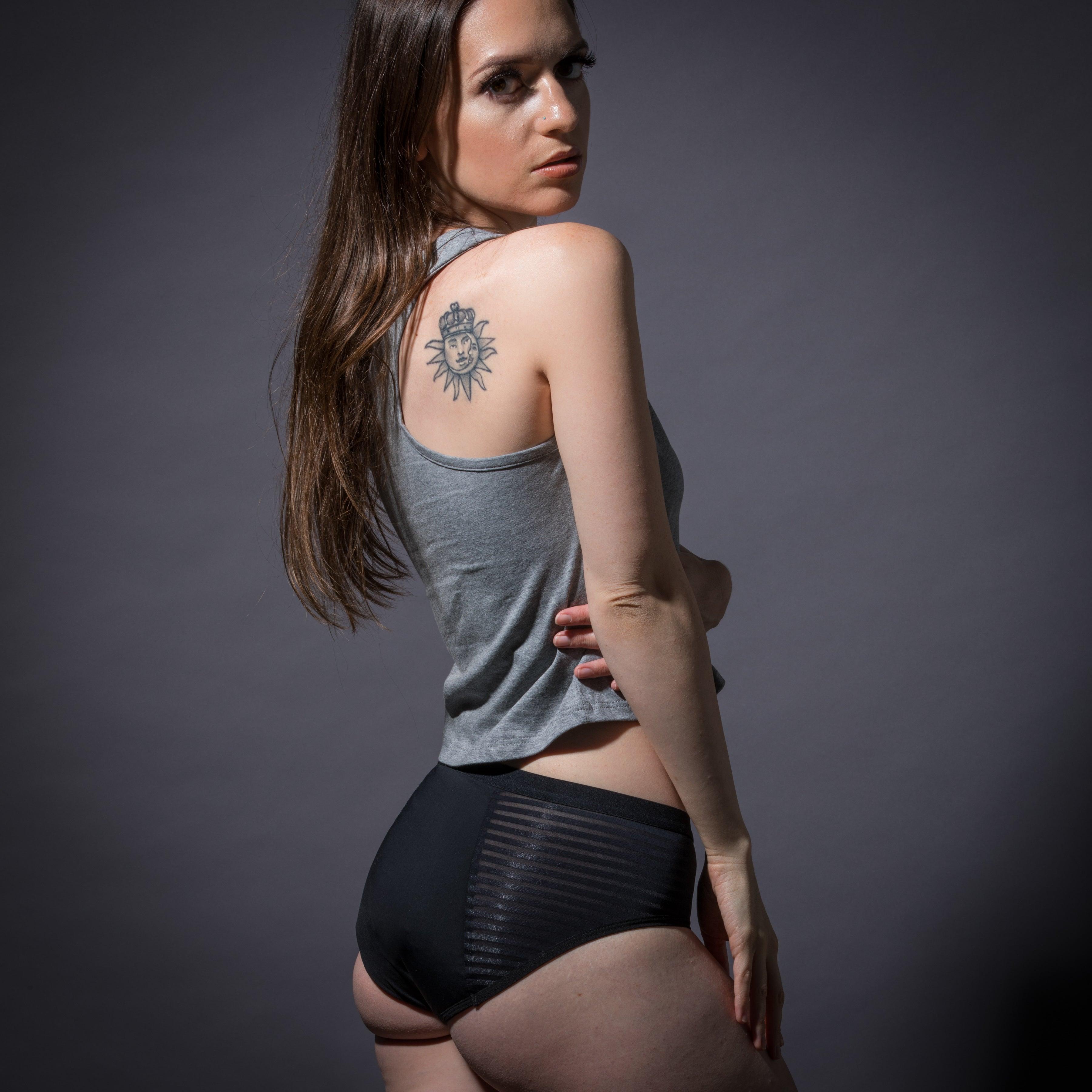 Woman wearing black leak-proof period underwear and a gray tank top, showing a sun tattoo on her shoulder.