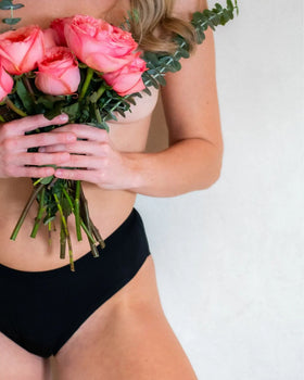 Close-up of a woman holding a bouquet of pink roses while wearing black Heralogie period underwear, symbolizing feminine empowerment and sustainable care.